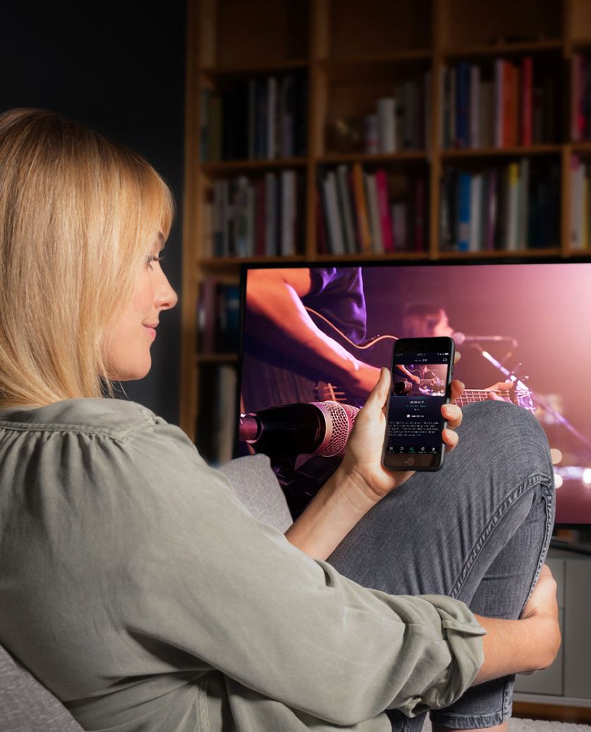 Frau auf dem Sofa mit Smartphone in der Hand. Im Hintergrund läuft der Fernseher