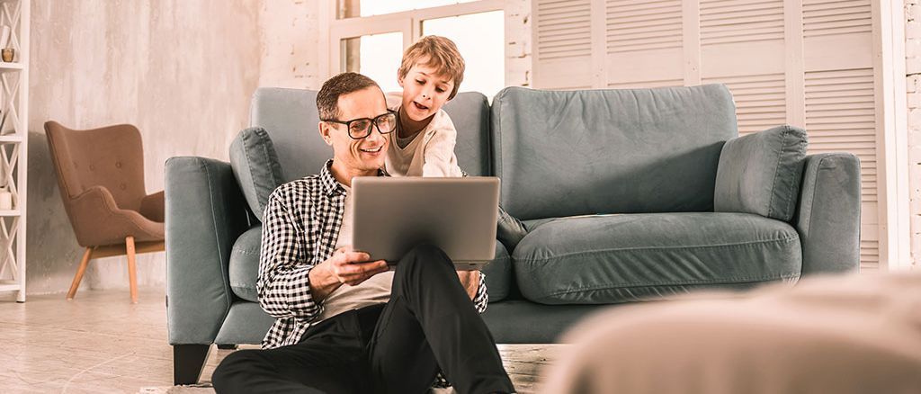 Mann mit Kind sitzend am Laptop vor dem Sofa